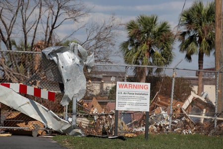 Debris litters Tyndall Air Force Base following Hurricane Michael on October 17, 2018 in Panama City, Florida. A sign in front of a fence with debris hanging off of it reads, "WARNING," "U.S. Air Force Installation." Palm trees and destroyed buildings are seen in the background beyond the fence