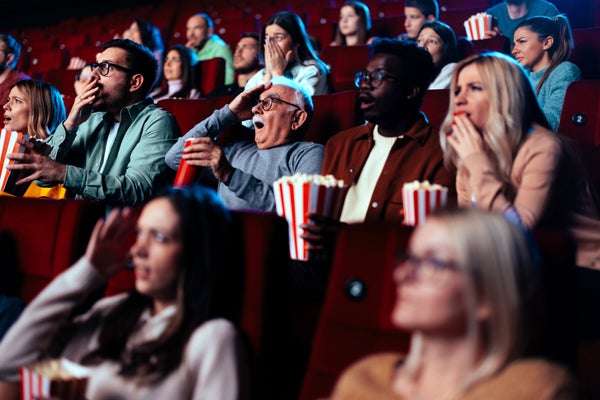A diverse audience is in the movie theater watching a scary movie and gesturing in fear.